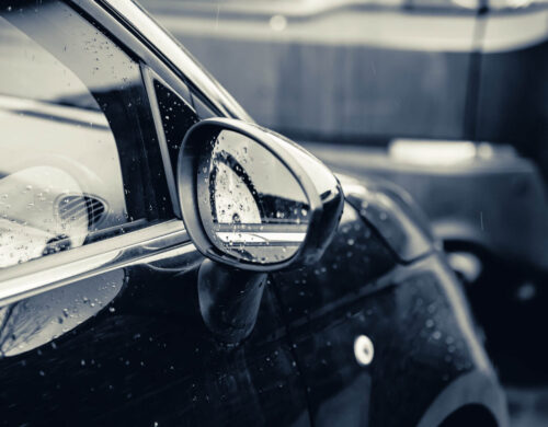 A closeup of a rearview mirror of a black car covered in raindrops with a blurry background